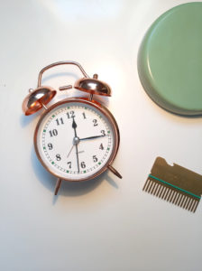 image of white table from above, a clock lies face up at 12:10pm. The the right is a gold credit card sized hair comb and in the top right corner, the base of a green lamp.