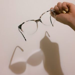 A hand is holding a pair of grey and white tortoise shell glasses. The lighting casts a shadow against a wall behind which is full, giving the illusion of being cast by a pair of sunglasses.