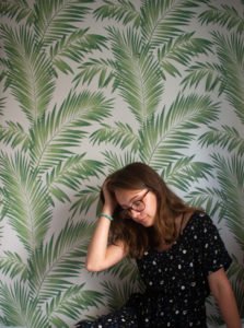 Eleanor sitting in her room, infront of a wall decorated in patterned palm leaf wallpaper.
