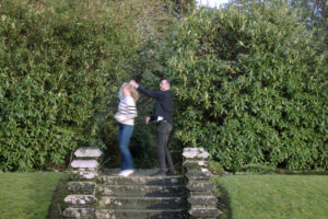 couple waltz in Loch Lomond walled garden, Scotland. they are spinning and holding hands infront of tall greenery. Some stone steps in front of them lead down towards the camera.