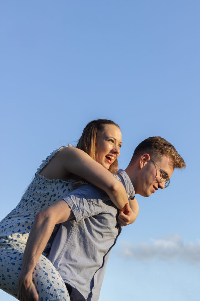 Fun and romantic engagement photo in London with a couple laughing and enjoying a piggyback ride against a bright blue sky. Capture the playful moments of your engagement with affordable photography by Eleanor Hyde at Distinctivemode in Edinburgh